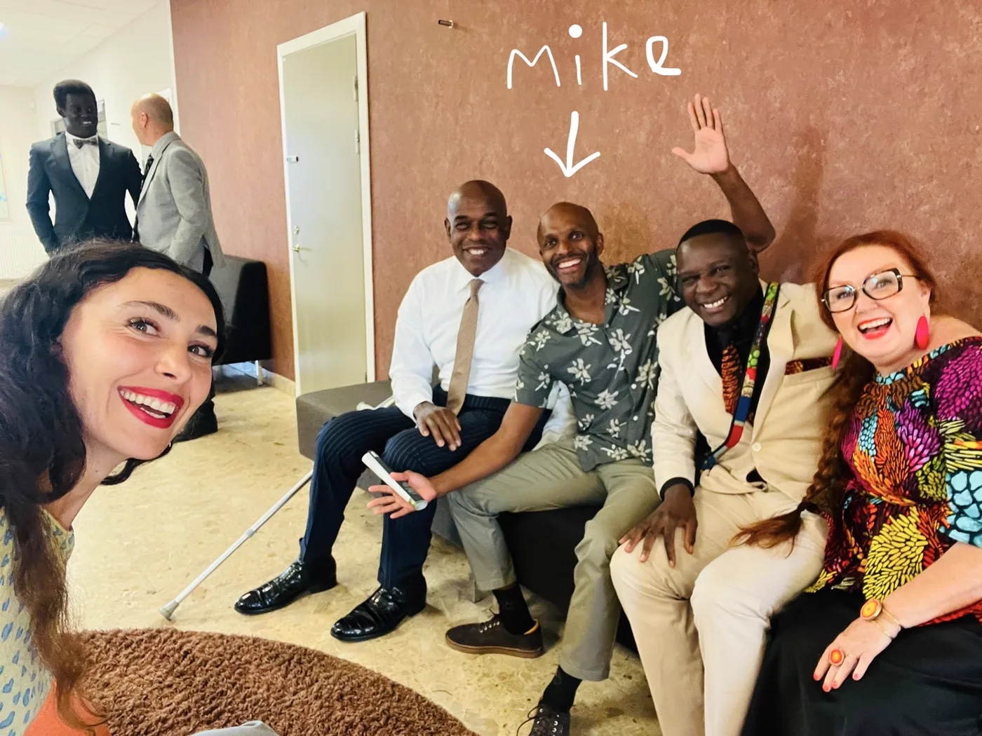 Michael laughing on a sofa with four friends at an event, wearing the olive shirt with cream flowers, a handwritten arrow above him labelled mike